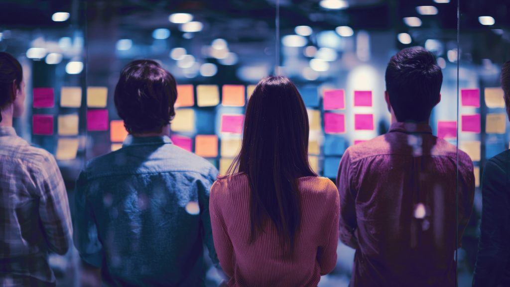 vier Personen von hinten schauen auf Post It Wand